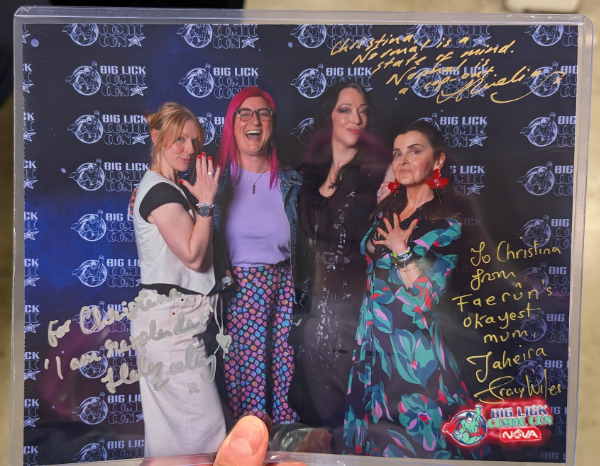 A commemorative photo of Tina, the author of this report, with three voice actors from the video game Baldur's Gate 3. From left to right is Helen Keeley, Tina, Amelia Tyler, and Tracy Wiles. They are standing in front of a dark blue backdrop and all are looking at the camera while posing. The photo is signed by the actors in shiny gold and silver marker.
