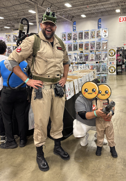 A very tall person in Ghostbusters cosplay smiling and standing next to a small child in a matching Ghostbusters cosplay. They are both posing to have their picture taken at a convention.