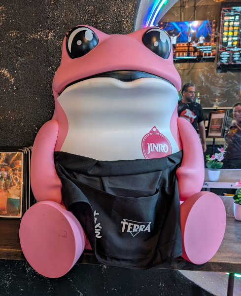 A photo of a large pink statue of the Jinro Soju Toad mascot in the lobby of a Korean-style barbecue restaurant. It is sitting on a counter and wearing a black apron.