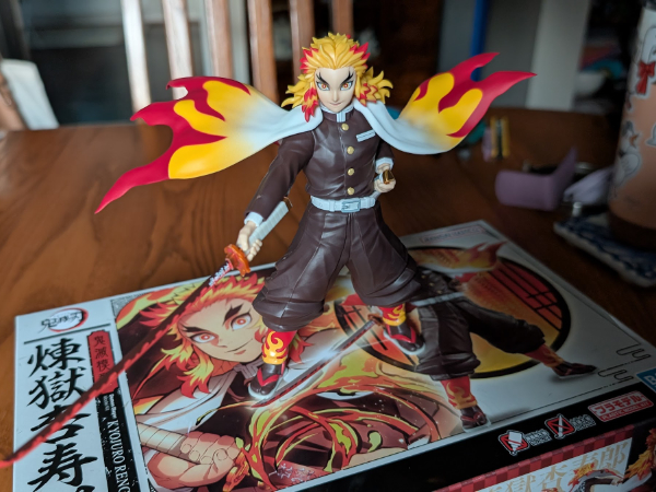 A photo of a plastic figurine of Rengoku from the anime and manga series Kimetsu no Yaiba. The figure depicts Rengoku posing with his sword unsheathed and at the ready, and his cape blowing in the wind.